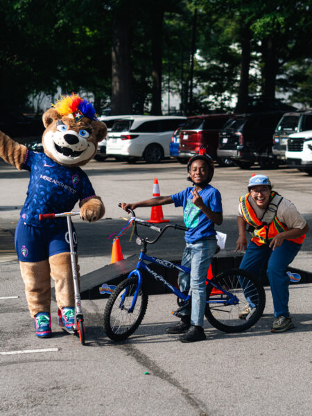 a soccer team mascot that is a lioness posing with a kid on their bike and a bike instructor